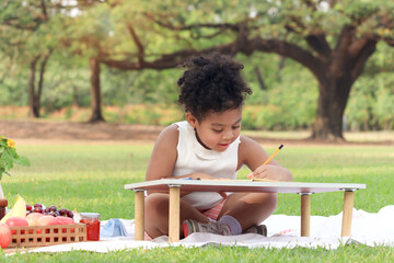 Happy smiling African little girl with black curly hair doing homework in outdoor garden. Child studying outside during having picnic in summer park. Cute kid study while sitting on mat on green grass