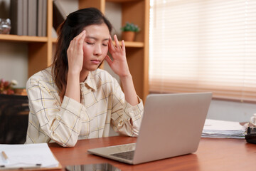 Asian professional with hand on forehead, eyes shut, exudes distress or deep thought at desk. Distraught woman leans on hand, laptop open, indicating mental strain or contemplation in quiet office.