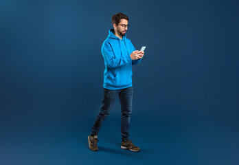Young Stylish Man Walking With Smartphone In Hands Over Blue Studio Background - obrazy, fototapety, plakaty