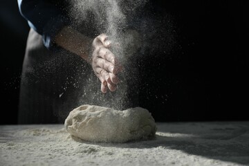 Making bread. Woman sprinkling flour over dough at table on dark background, closeup. Space for text