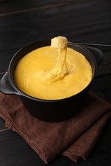 Dipping piece of ham into fondue pot with melted cheese on black wooden table
