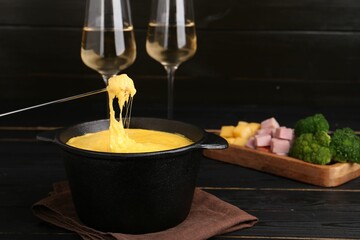 Dipping piece of ham into fondue pot with melted cheese on black wooden table