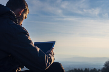Young man in jacket using tablet. Landscape with distant hill at sunrise, copy space sky