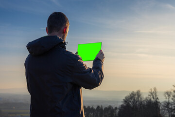 Young man in jacket using tablet with green screen. Landscape with distant hill at sunrise, copy space sky