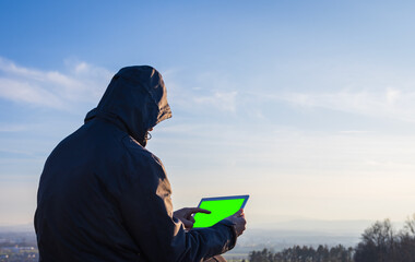 Young man in jacket using tablet, showing with finger on green screen. Landscape with distant hill at sunrise, copy space sky
