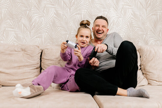 A father spends time with his little daughter, sitting on the sofa and playing a game console. Weekend activities, free time, home entertainment and video games concept.