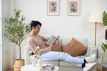 Happy asian man sitting on couch  reading a book in living room at home, relax time and lifestyle concept