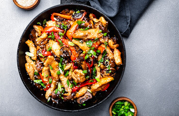 Asian stir fry chicken  slices with red paprika, mushrooms, chives and sesame seeds in frying pan. Gray kitchen table background, top view