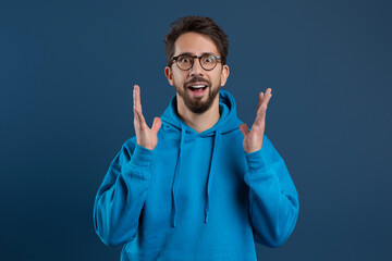 Enthusiastic young man with beard and glasses expressing astonishment with raised hands
