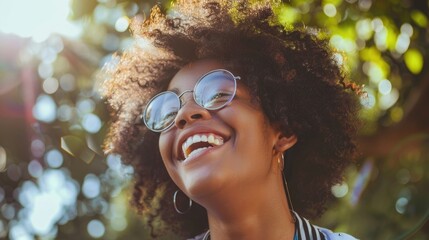 A joyful woman with curly hair laughing out loud, wearing glasses, and soaking up the sun among lush greenery. - obrazy, fototapety, plakaty