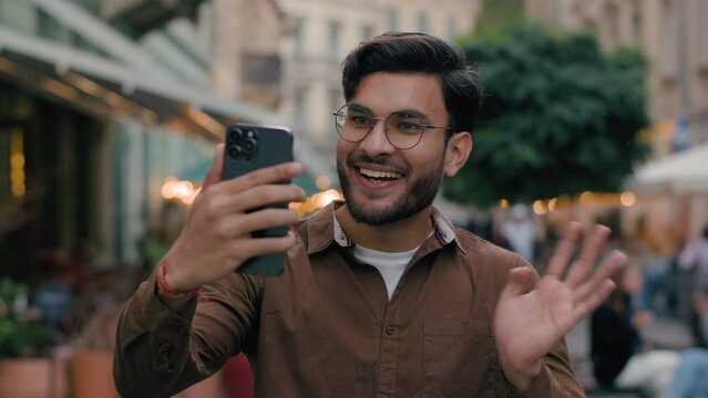 Happy smiling Indian Arabian ethnic man male student gen z guy blogger using modern cellphone recording video travelling blog mobile phone waving hand thumbs up social media outside abroad city street