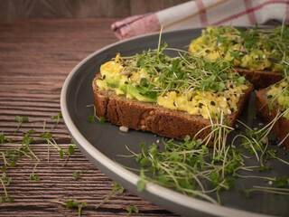 A snack of black bread with avocado and arugula sprouts on a gray plate on a wooden table. The concept of proper nutrition, low-calorie snacks and weight loss