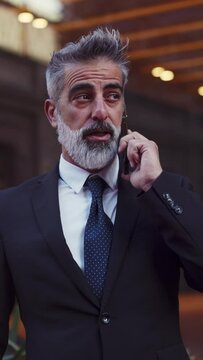 Mature businessman in expensive suit discussing deals over a mobile call, standing against urban backdrop. Successful male executive conversate work deals via smartphone near office. Vertical Format
