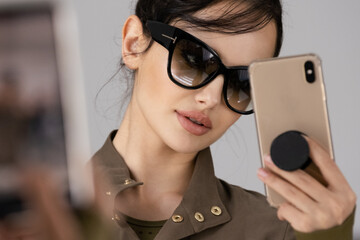 Portrait of a Girl with smartphone.  Stunning girl in stylish black sunglasses takes a selfie. Beautiful brunette woman  in sunglasses poses in front of a mirror with cell phone. - 755889385