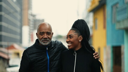 African American Mature father walks forward with arm around teenage daughter in urban street...