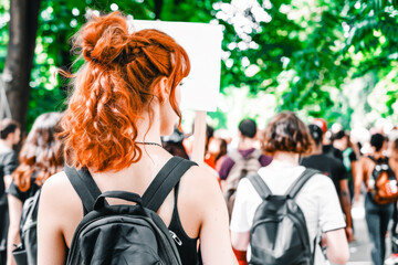 Rear view of a young copper-haired woman with backpack holding a blank sign in the middle of a crowd demonstrating in the street.