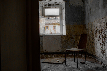 Ein einsamer Stuhl in einem zerstörten, maroden, kaputten, leeren Raum mit offenem Fenster und der Aussicht auf ein altes Gebäude mit vergitterten Fenstern