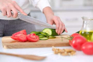 Woman cutting fresh cucumber and tomatoes at table in a light kitchen, closeup