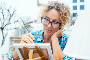 Picture of serious concentrated adult Caucasian female artist sitting at desk with painting accessories, looking the canvas and paint enjoying hobby and indoor leisure activity alone. Mature lady home