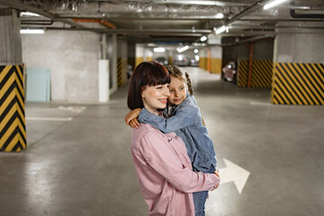 Fototapeta premium Portrait of Caucasian mother holding her preschool daughter in hands having fun in underground parking lot. Happy family in casual clothes walking to their car before an exciting trip.