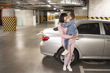 Attractive Caucasian mother holding preschool daughter in hands on background of modern car in...