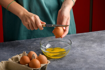 A woman's hand cracking an egg with knife into a clear glass bowl in the gray kitchen table, the process of cooking, hands holding the eggshells, Lifestyle, close up