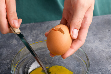 A cracked egg in the hand, close up. A woman's hand cracking an egg with knife into a clear glass bowl in the gray kitchen table, the process of cooking, Lifestyle, close up