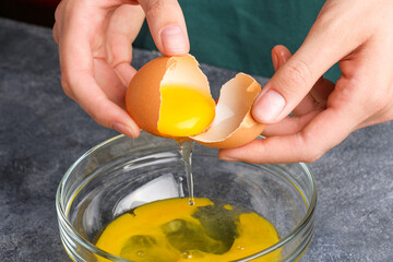 Housewife hands cracking fresh egg, yolk and white dropping in a bowl. A woman's hand cracking an egg with knife into a clear glass bowl in the gray kitchen table, the process of cooking, close up