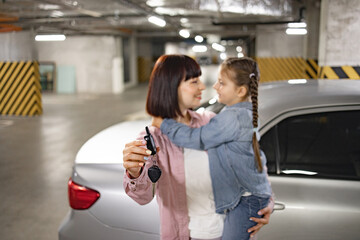 Selective focus of hand of mother and daughter prepared keys to open car for weekend trip. Happy Caucasian woman holding female kid and car keys in underground parking lot.