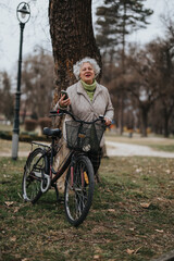 Mature woman with curly hair and a bright smile stands with her bicycle in a tranquil park setting, embodying an active lifestyle and joy.