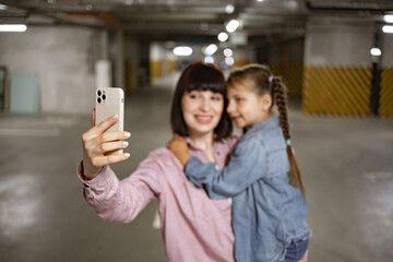 Happy family in casual clothes having video call using cell phone before exciting trip. Caucasian mother holding preschool daughter in hands making selfie with smartphone in underground parking lot.