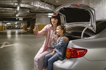 Caucasian family having video call using cell phone sitting on the trunk of car before exciting...