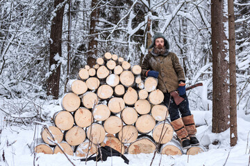 A forester prepares firewood in a winter snowy forest and stands with an ax near stacked stumps....