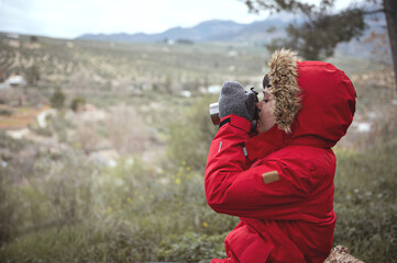 Teenage boy in red parka, drinking hot tea from a steel thermos mug, sitting in the forest against mountains background
