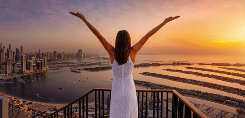 A happy woman looks at the panoramic view of the Dubai Marina district and the Palm island during golden sunset time, UAE