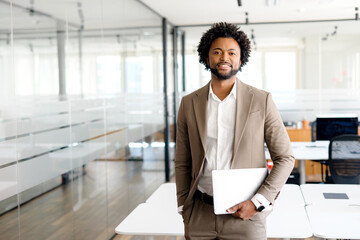 Fototapeta premium A cheerful entrepreneur holds his laptop while standing, his posture exuding confidence and professionalism within a sleek office space. Mobility in business, cloud computing, on-the-go professional