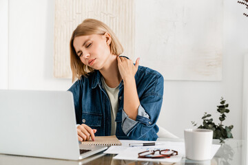 Tired young woman rubbing neck having neck pain while working on laptop sitting at the desk. Office...
