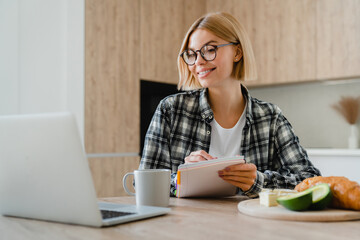 Happy young woman taking notes while working on laptop at home kitchen. Female freelancer...