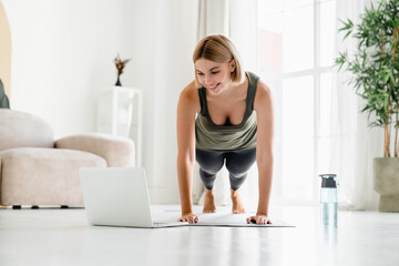 Young caucasian fit woman standing in plank position while watching online training on laptop. Female athlete improving stamina endurance