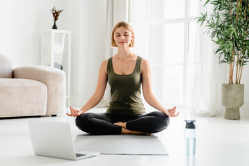 Fototapeta na wymiar Young caucasian fit female sitting in lotus position during online training on laptop. Home yoga exercises, meditation and serenity concept