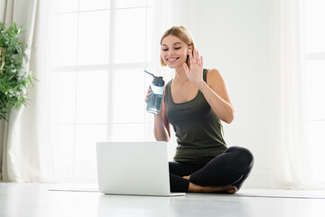 Happy young female athlete in sporty clothes sitting on fitness mat while talking on videocall on laptop. Online training, lesson of fitness. Social distancing concept