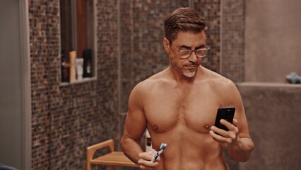 Mature man multitasking by brushing his teeth and browsing his phone in a modern bathroom. Portrait of muscular fit aged male. - 755944361