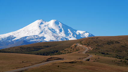 A narrow winding highway along the mountain plateau to the snowy Elbrus against the blue sky. Copy space.