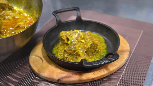 Chef in black glove in kitchen prepares Georgian dish Ostri, in cast iron pan on wooden board on table in restaurant . Adds meat in sauce to hot scoop.