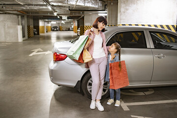 Attractive female family dressed in casual attire, sharing joyful moment of togetherness. Underground parking lot, Caucasian mother and little daughter with bags with clothes bought in shopping mall.