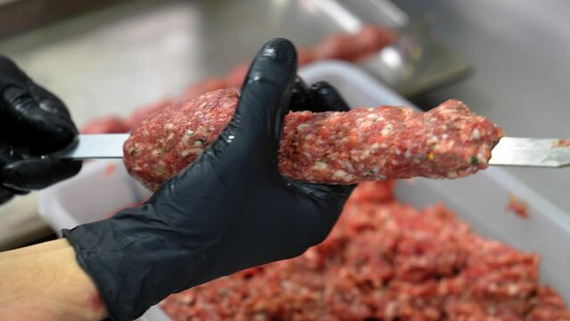 Chef sticks forcemeat on skewer and forming it for cooking lula kebab on grill at commercial kitchen. Close-up hands in black gloves