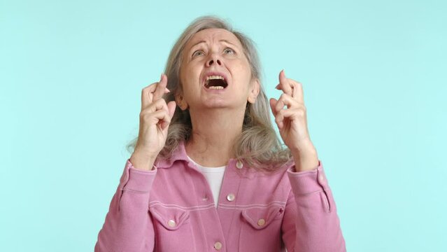 Optimistic elderly woman with fingers crossed in a gesture of hope and anticipation, wearing a pink shirt, looking for good luck or a positive result. Camera 8K RAW. 