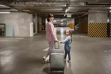 Youthful lady with her child clutching travel suitcase searching for vehicle before starting journey. Happy mom and small daughter, attired in informal wear, stroll through subterranean parking area. - obrazy, fototapety, plakaty