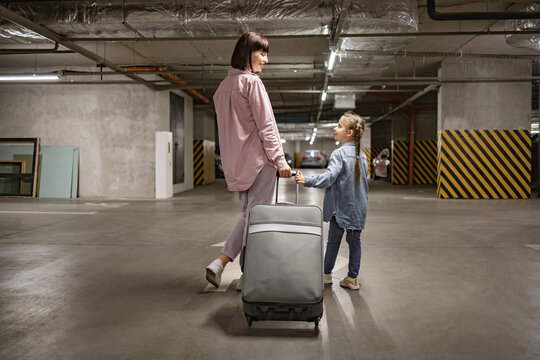 Dressed in informal attire, Caucasian mom and small daughter walking through underground parking lot. Attractive lady with her child and travel suitcase, searches for their car before starting trip.
