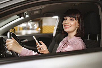 Fototapeta na wymiar Attractive female typing on phone inside gray vehicle on underground parking lot. Young confident Caucasian woman in casual clothes sitting behind wheel of modern car with smartphone.